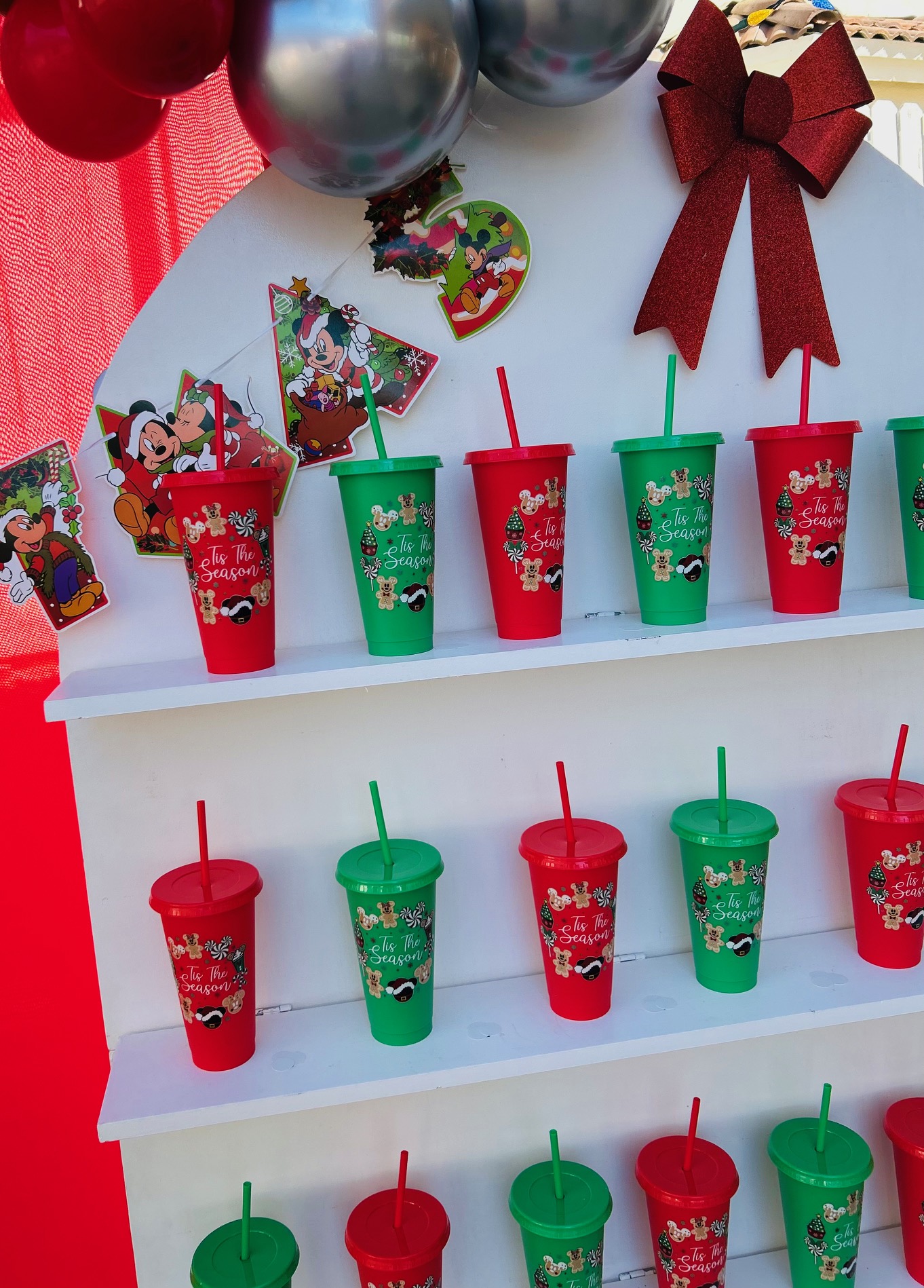 Holiday-themed curated favor wall with red and green custom cups, Mickey decor and festive bow